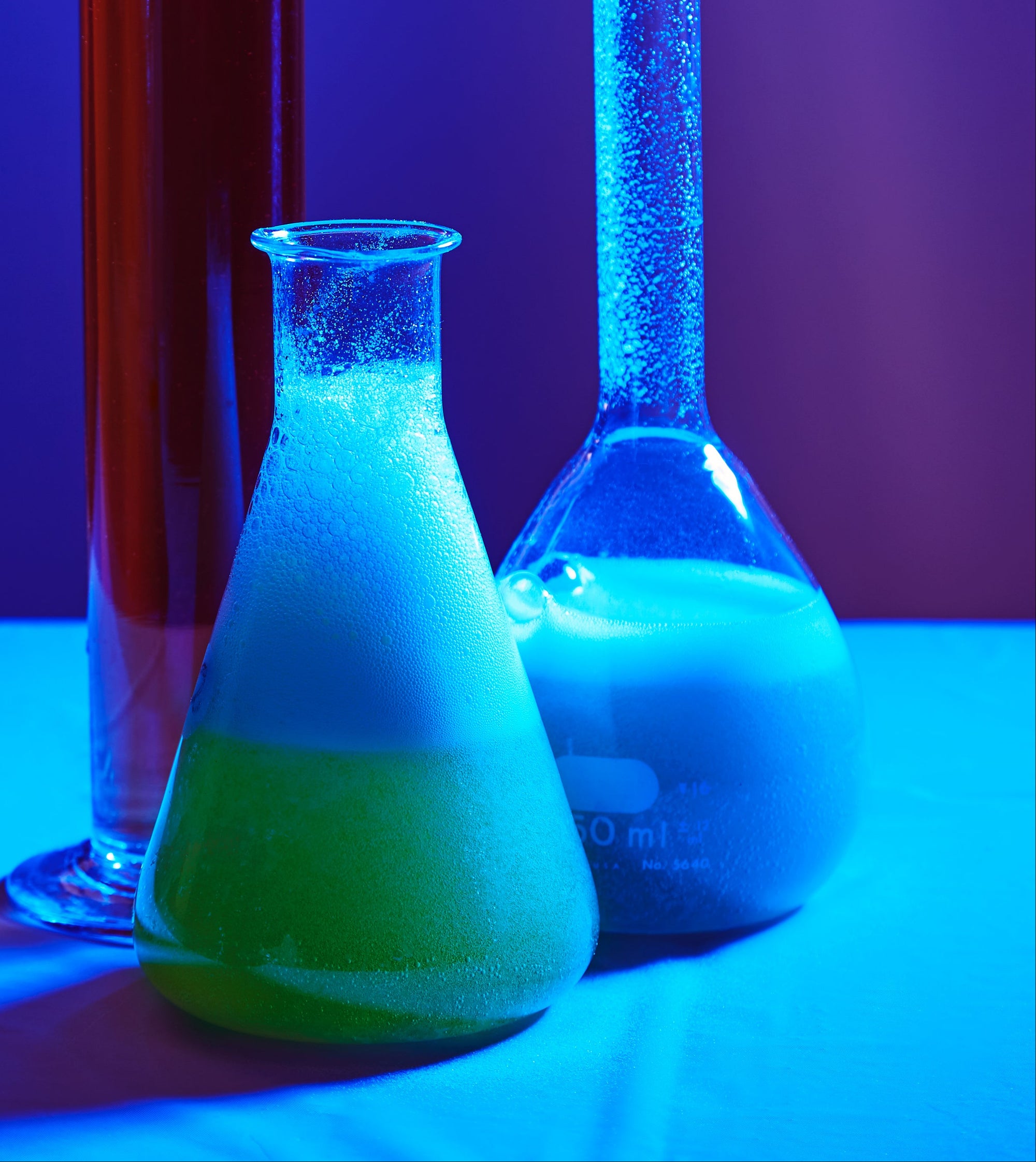 Two beakers with glowing green and blue liquids on a reflective surface under UV light.